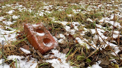 Red brick partially covered in snow and surrounded by fallen leaves and grass, showcasing the contrast of nature and man-made materials in a winter landscape
