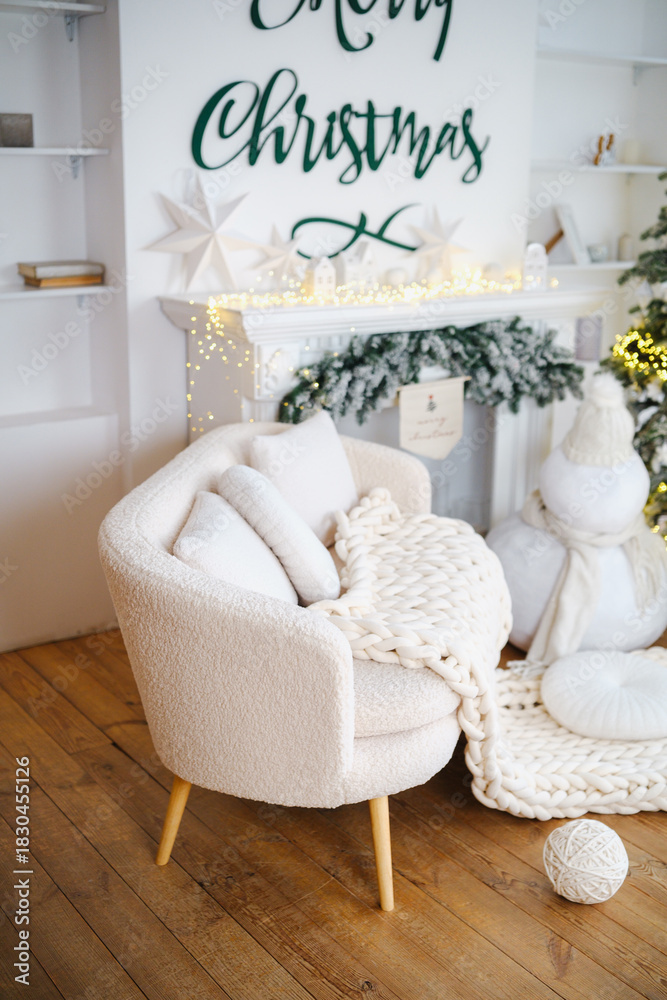 Naklejka premium Cozy cream armchair with warm knit blanket near decorated fireplace and Christmas tree