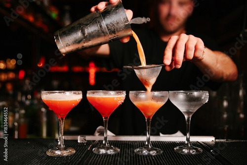 Professional mixologist pouring and straining a craft cocktail from a metal shaker into a row of coupe glasses on a dark bar counter