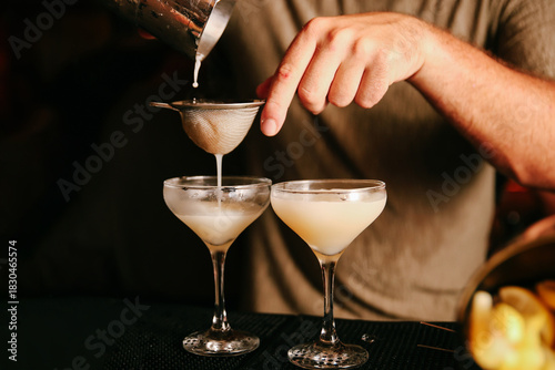 Photos Close-up of a mixologist straining a clear cocktail into coupe glasses highlight