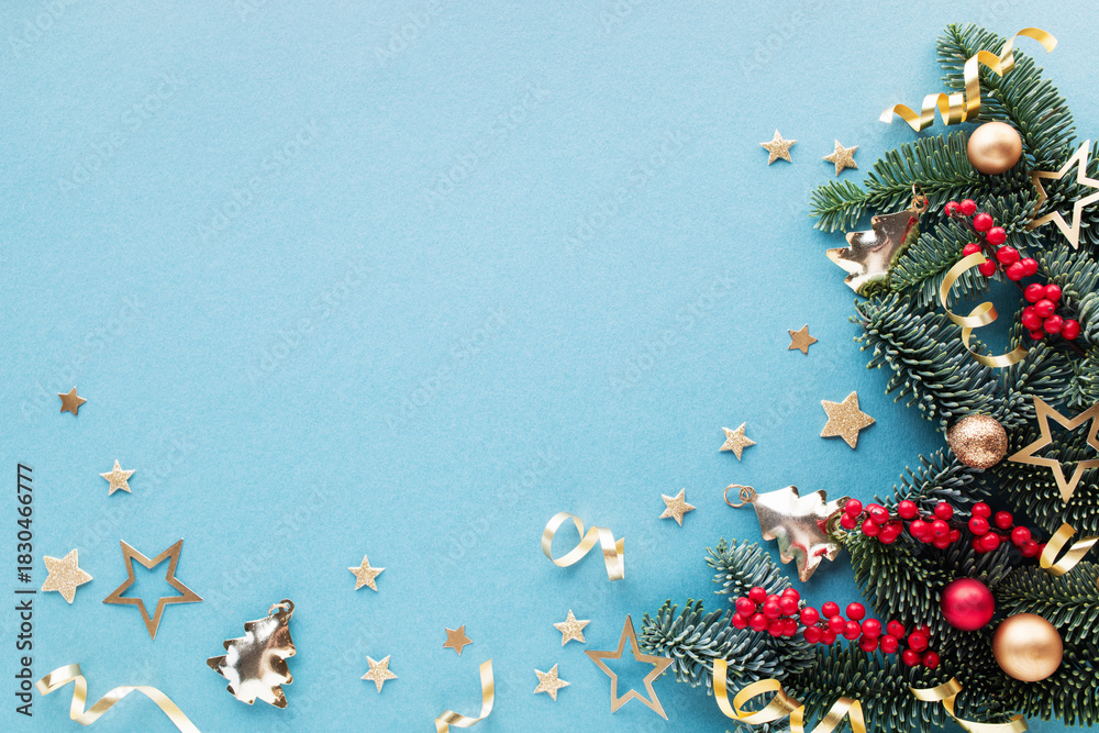 Obraz premium Festive Christmas background or greeting card with green pine branches, red berries and shiny golden decorations on blue table top view, flat lay.