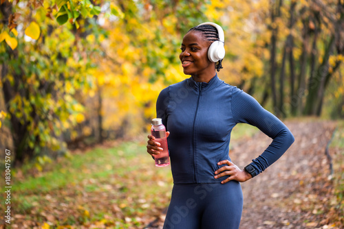 A runner stands on a wooded path, looking relaxed and happy, with vibrant yellow and green leaves creating a lively autumn atmosphere around her.