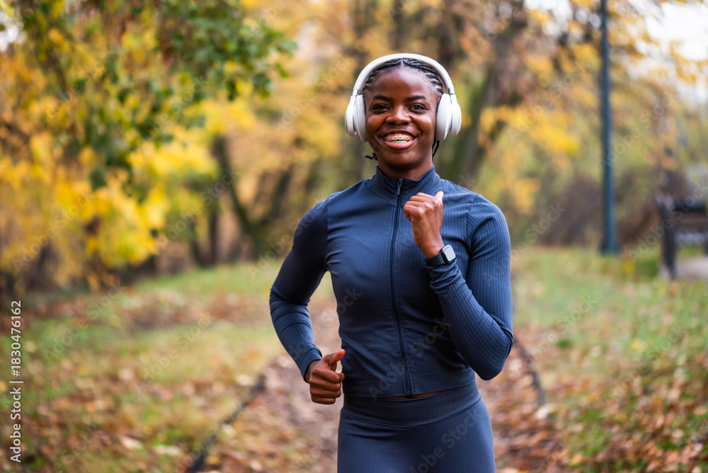 Obraz premium Cheerful woman jogs along a leaf-covered forest trail, surrounded by warm autumn colors and tall trees as she enjoys an outdoor run.
