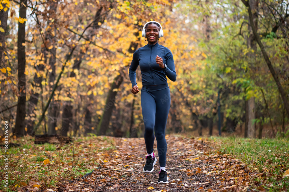 Fototapeta premium Happy black woman runs on running trail of fallen leaves, with golden and green trees creating a serene, seasonal backdrop.