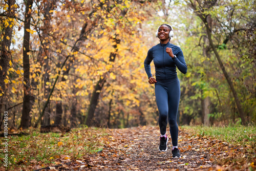 A smiling black woman runs on running trail of fallen leaves, with golden and green trees creating a serene, seasonal backdrop.