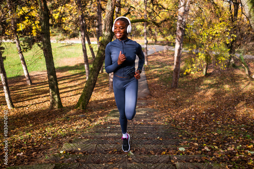 In a forested park with vibrant fall colors, a smiling runner in fitted sportswear climbs outdoor steps while listening to music.
