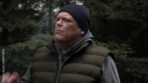 A middle-aged man in outdoor attire smoking a cigarette, with his face in profile against a blurred dark green forest background. The scene depicts addiction, bad habits, and outdoor break time.