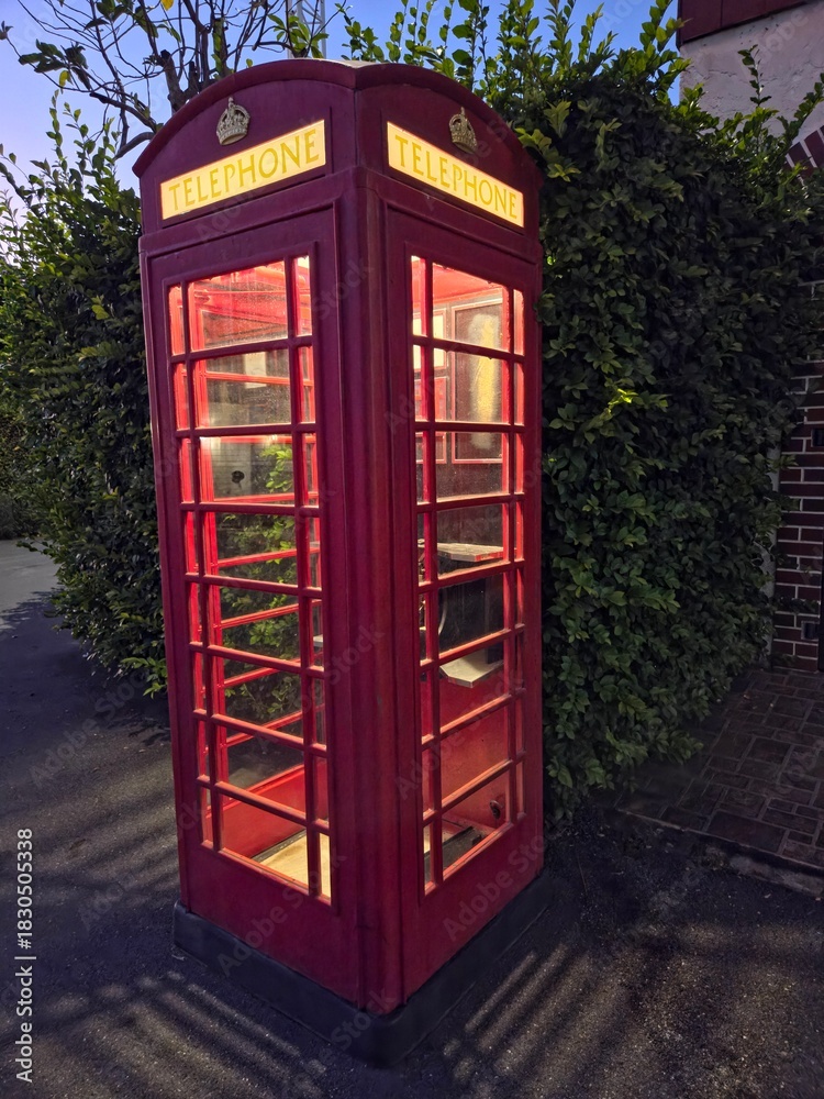 Obraz premium A classic red British telephone booth (kiosk) illuminated from within, standing on a dark pavement next to a dense green hedge and a brick wall, likely taken at twilight or night.