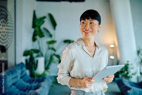 Confident Asian businesswoman holding a digital tablet with a happy expression, standing in a contemporary office setting
