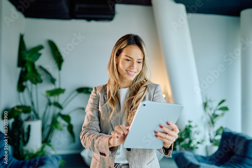 Smiling young businesswoman using a digital tablet for work, standing in a contemporary office space with plants