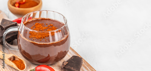 Spicy hot chocolate with chili pepper, cocoa powder and dark chocolate pieces served in a glass mug on a light wooden surface, captured in a flat lay composition with wide copy space for text