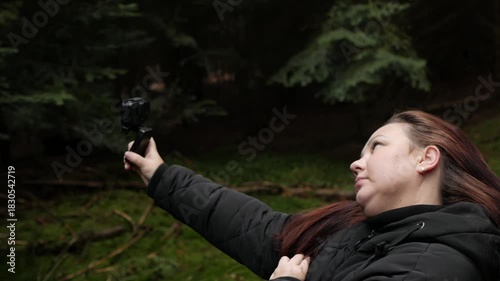 A side-profile shot of a female blogger or hiker in a dark jacket holding an action camera up to film herself against a dark, dense pine forest background. 