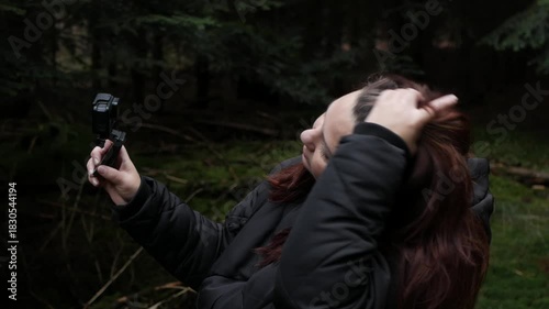 A side-profile shot of a female blogger or hiker in a dark jacket holding an action camera up to film herself against a dark, dense pine forest background. 