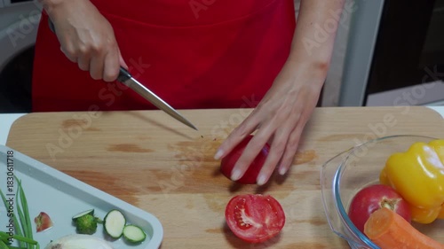 Female hands slice a fresh red tomato on a wooden cutting board using a knife. The cook prepares ingredients for a healthy salad in the kitchen. A bowl of colorful vegetables sits nearby.