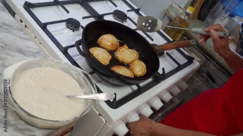 A woman fries golden pancakes in a black pan on a white gas stove. She adjusts the heat knob while holding a spatula. A bowl of fresh batter sits on the kitchen counter nearby.