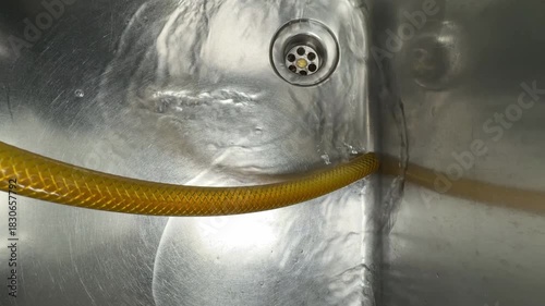 A close-up view of a stainless steel sink with water flowing around a yellow flexible hose. The metallic surface reflects light, creating abstract patterns and textures.