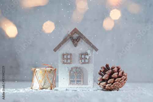 Festive winter scene with a white miniature Christmas house, snow covered pine cone and golden drum decoration on snowy surface with soft bokeh lights in the background.