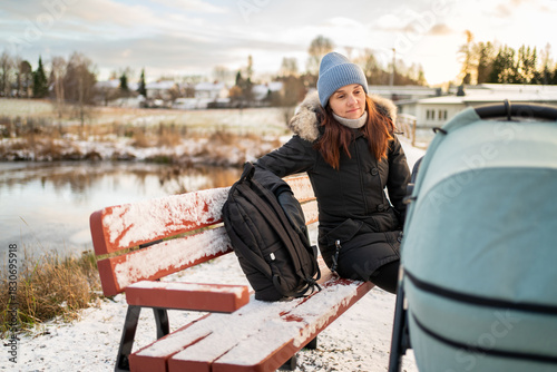 Mother and stroller. Baby sleeping in pram or buggy. Tired woman sitting on park bench in winter. Child carriage and pushchair.