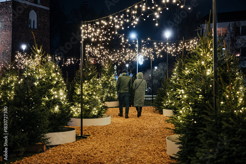 People walk through a festive outdoor Christmas tree display adorned with glowing lights and illuminated bird decorations in an urban night setting