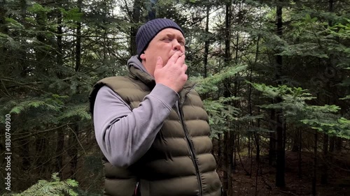 A middle-aged man in outdoor attire smoking a cigarette, with his face in profile against a blurred dark green forest background. The scene depicts addiction, bad habits, and outdoor break time.