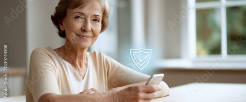 Smiling elderly woman holding smartphone with digital shield icon representing security and protection in bright home interior