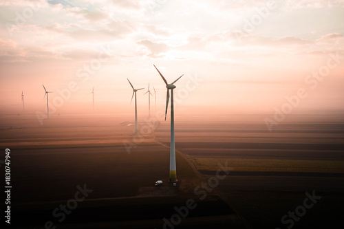 Wind turbines rising above soft morning fog, illuminated by warm golden sunrise tones. The calm landscape blends mist, light and motion, creating a serene and atmospheric view of modern renewable ener