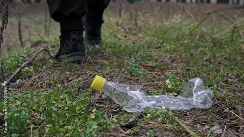 A male foot wearing a black sneaker steps firmly on a discarded plastic bottle lying in the grass. The action demonstrates the crushing of waste and the prevalence of pollution in green spaces.