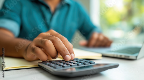 Close-up of a person's hand using a calculator on a desk next to a laptop, managing personal finances or business calculations.