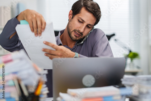 A busy businessman handles document work and takes a smartphone call in a messy office while feeling overworked
