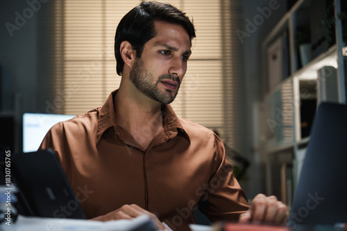 A businessman works at his laptop at night while reviewing documents and managing an overtime workload.