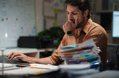 A tired businessman lets out a sleepy yawn while working through paperwork at night in an overworked office.
