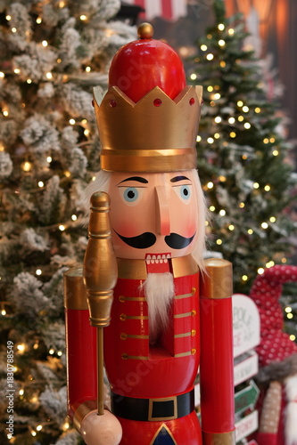 Close-up of a red and gold Nutcracker soldier with a crown, surrounded by frosted Christmas trees and holiday lights.