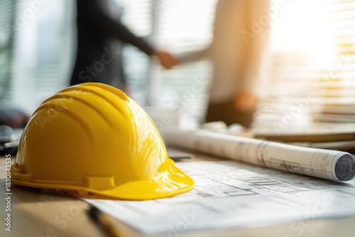 Yellow hard hat rests on blueprints with blurred handshake background.