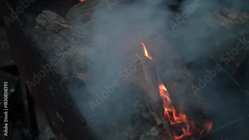 A close-up of burning wood in a barbecue grill. The smoldering embers, crackling wood, and rising flames create a warm, rustic atmosphere.