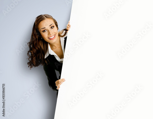 Happy smiling cute brunette haired woman peep out show pointing advertise blank white banner signboard with empty sign text free space, isolated grey gray background. Business ad concept
