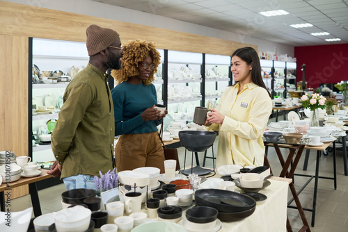 Young Black man and young Black woman standing together listening to young Caucasian woman sales assistant explaining ceramic tableware in modern store, surrounded by dishes and bowls