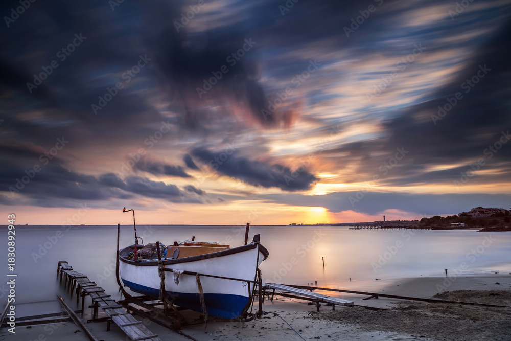 Obraz premium Long-exposure coastal scene at sunset featuring an old wooden fishing boat resting on a sandy shore beside wooden docks. Ravda, Burgas, Bulgaria