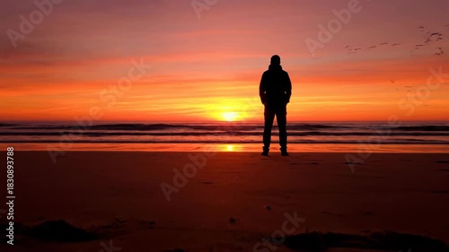 Person silhouette stands on beach against vibrant sunset ocean view