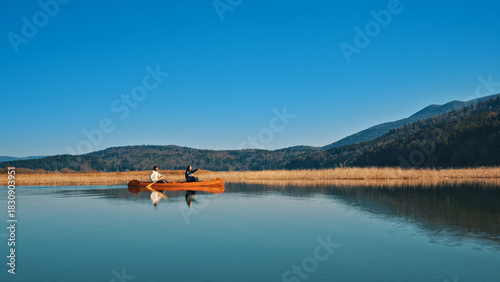 A couple paddles a canoe on a serene lake, surrounded by rolling hills. The clear blue sky and calm water reflect their peaceful activity, capturing a moment of connection and tranquility in nature.
