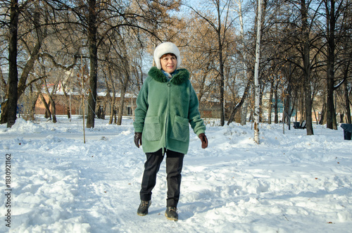 Mature woman in warm winter coat and white fur hat walks away down snowy park path, enjoying a peaceful cold day outdoors