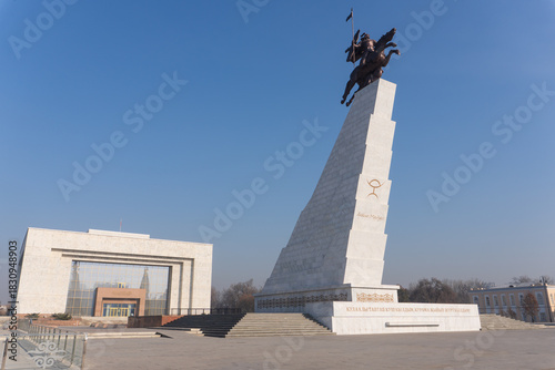 Wide view of Ala-Too Square featuring Manas monument on pedestal right and National History Museum white building left paved plaza clear blue sky. Bishkek, Kyrgyzstan.