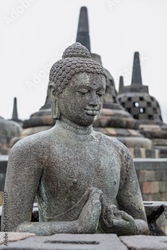 Yogyakarta Indonesia 12 11 2025 – Stone Buddha in the foreground at Borobudur Temple with iconic stupas rising behind under the open sky.