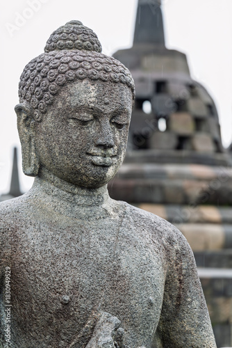 Yogyakarta Indonesia 12 11 2025 – Stone Buddha in the foreground at Borobudur Temple with iconic stupas rising behind under the open sky.
