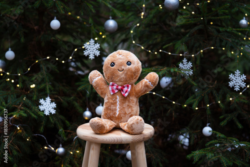 A cute gingerbread plush toy standing in front of a beautifully decorated Christmas tree, surrounded by warm holiday lights and festive ornaments. The cozy Christmas atmosphere.