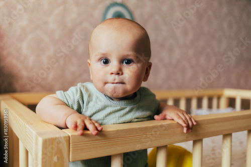 Cute baby sitting on a soft rug in a nursery next to a wooden crib. The child is holding a plush toy and looking at the camera with a gentle smile. Warm home lighting, natural tones, and a cozy atmosp