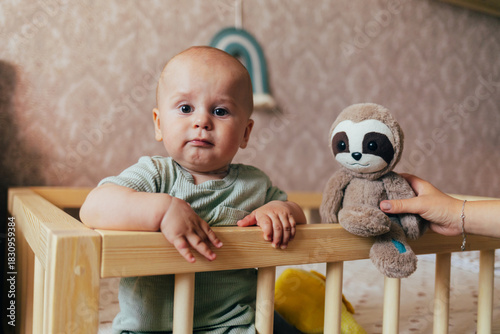 Cute baby sitting on a soft rug in a nursery next to a wooden crib. The child is holding a plush toy and looking at the camera with a gentle smile. Warm home lighting, natural tones, and a cozy atmosp