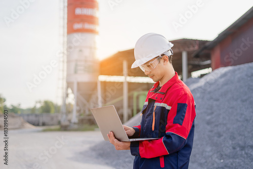 An engineer standing at industrial material storage yard checking data plans on laptop. Project management quality control. Large cement silo and pile of materials in background.