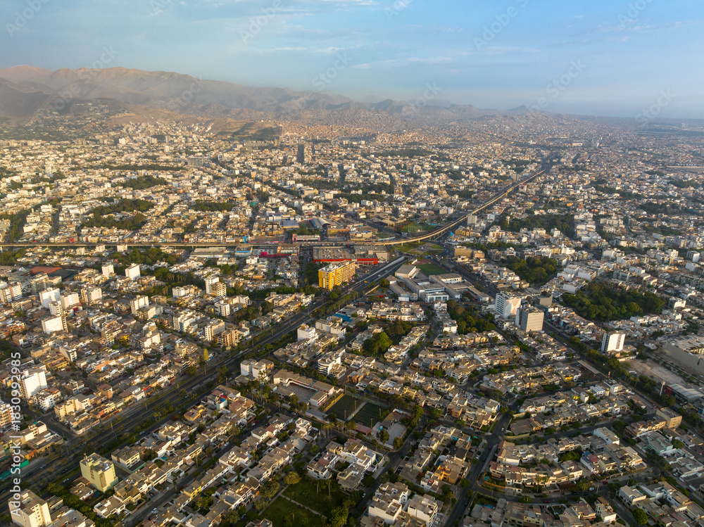 Fototapeta premium Aerial view of Ovalo Higuereta in Lima, Peru, connecting Surco and Miraflores districts, featuring the Metro train line, urban density, and mountains in the background during sunset.