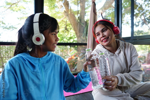 A close up shot of two females clinking water bottles after a yoga session. Both are wearing headphones and smiling at each other.