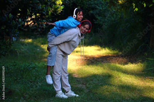 The mother is giving her daughter a piggyback ride while both are smiling and posing like they are flying in the park.